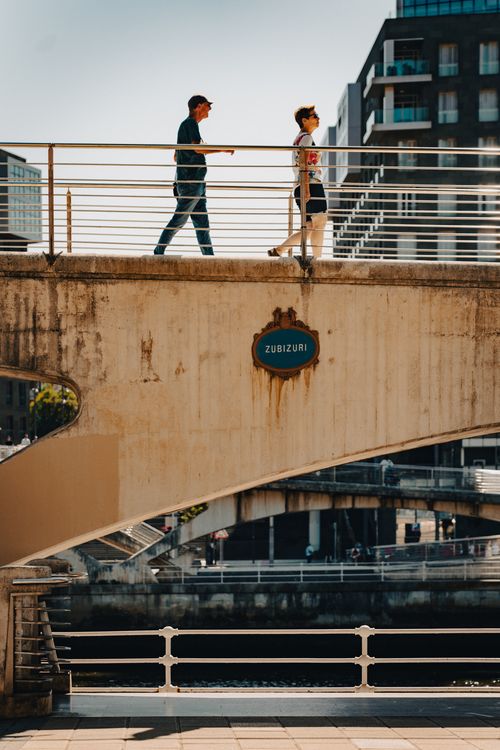 Free Two people walking on Zubizuri Bridge, Bilbao Spain, amidst urban backdrop on a sunny day. Stock Photo