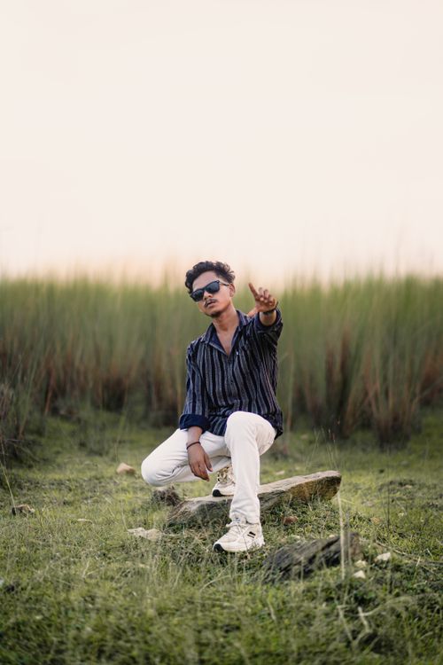 Free Young man in sunglasses sitting on stone in open grass field under bright sky. Stock Photo
