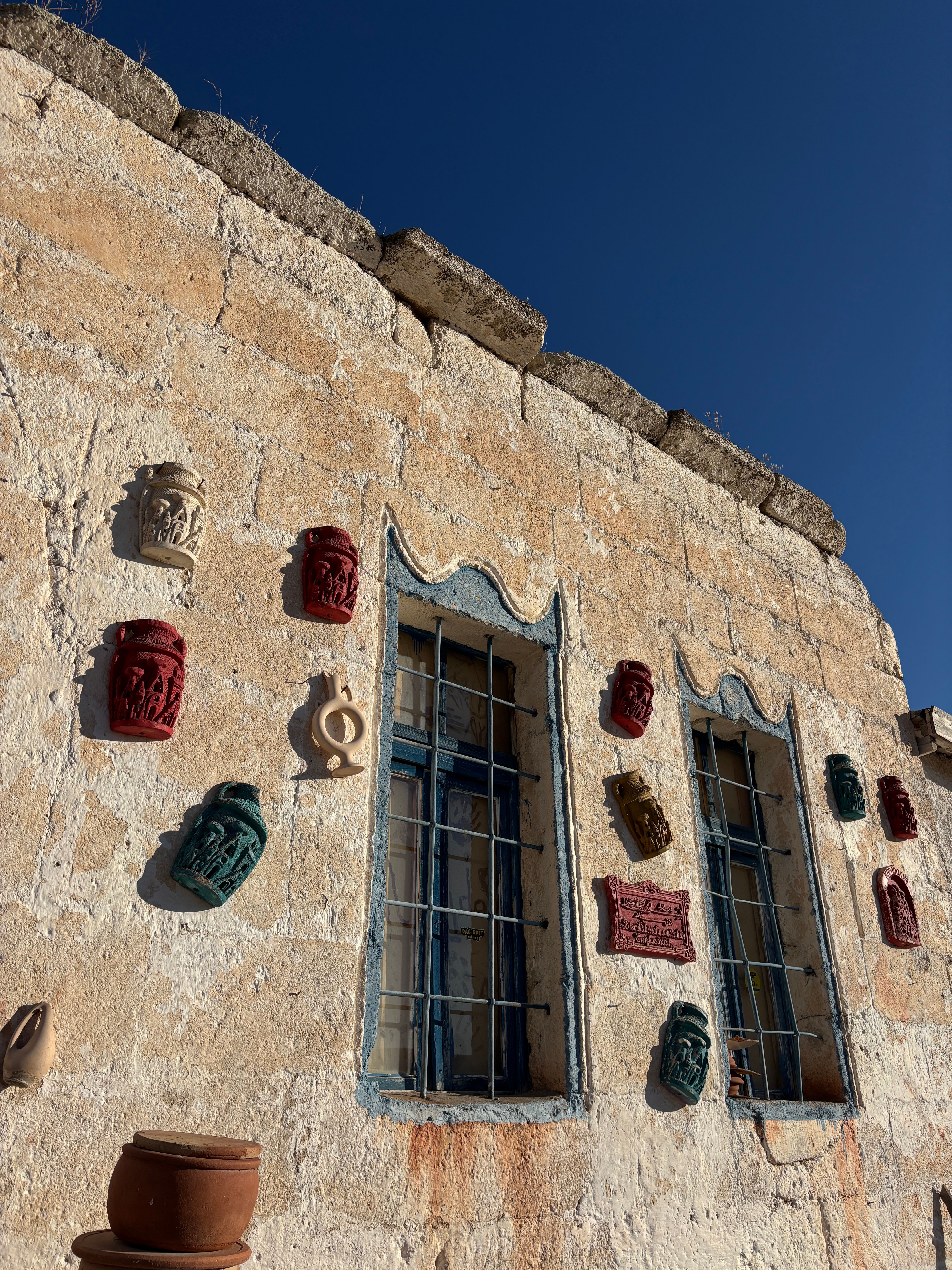 Fotomural Muro de piedra bañado por el sol en Capadocia, adornado con coloridas máscaras decorativas y ventanas rústicas.