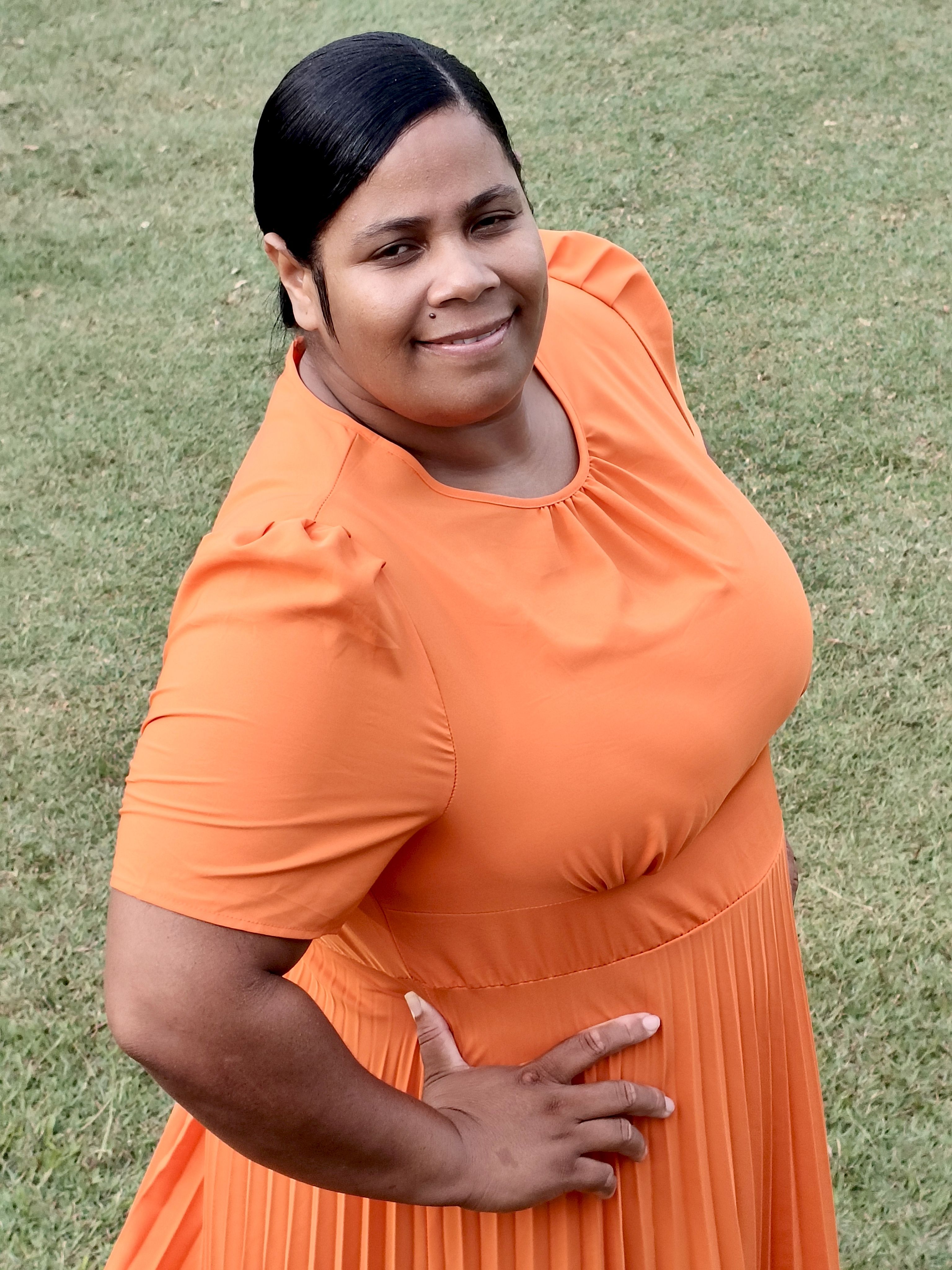 Portrait of a smiling woman in an orange dress standing on grass outside.