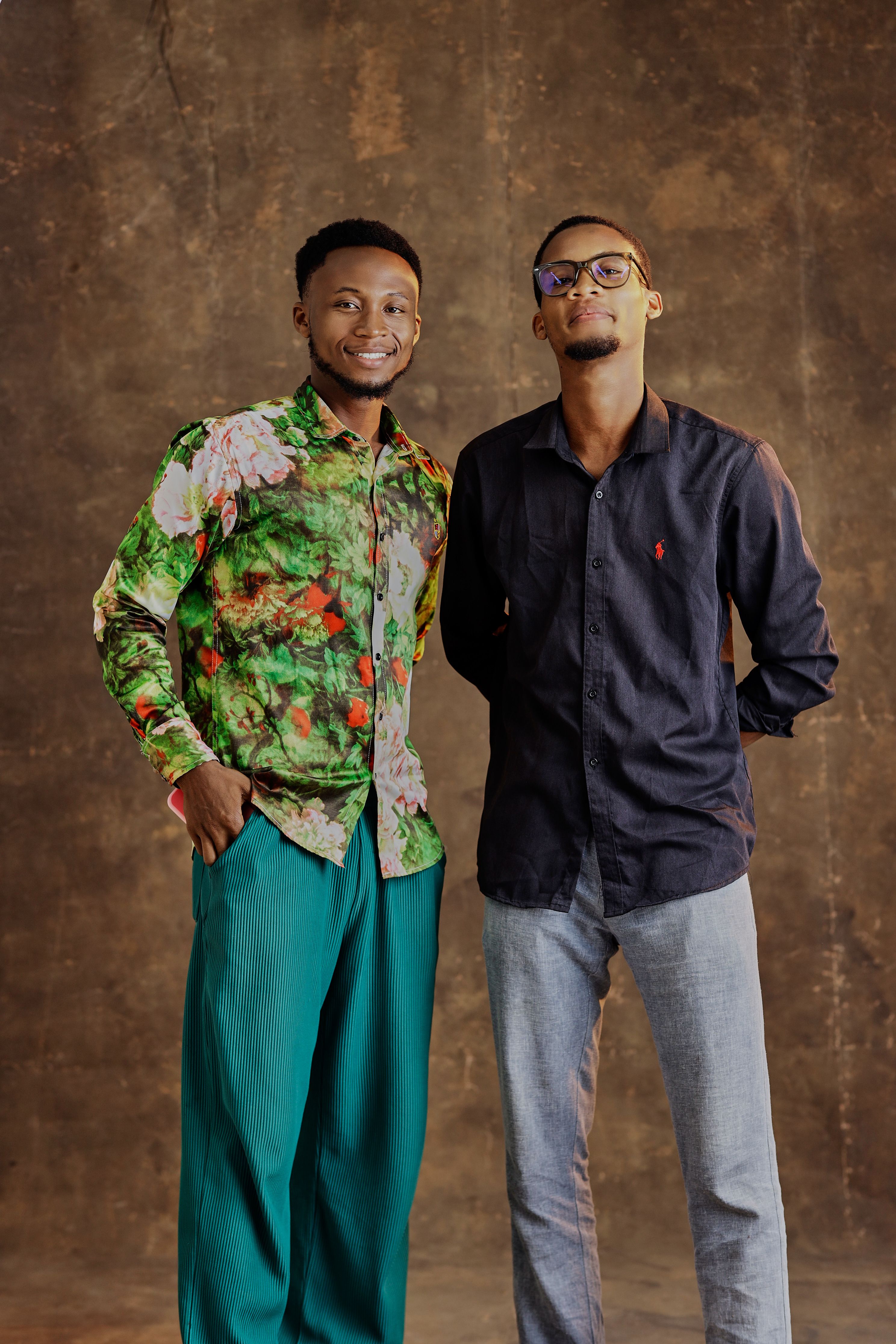 Two young men wearing fashionable clothing pose indoors against a textured background.