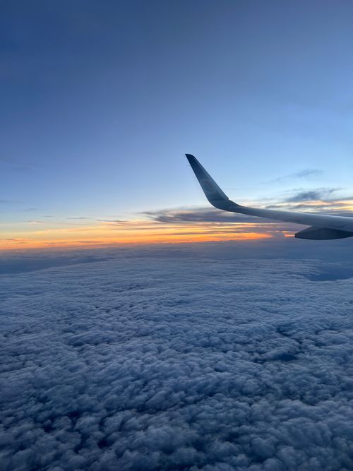 Free A serene aerial view of an airplane wing flying above clouds during sunrise, with clear skies and vibrant colors. Stock Photo