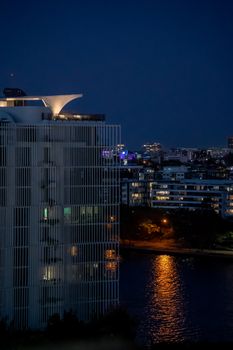 Urban skyline at night reflecting on a river, featuring modern architecture.