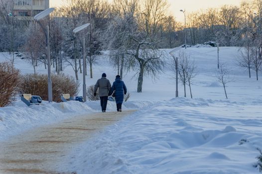 A serene winter scene in Minsk showing two people walking on a snowy path during dusk.