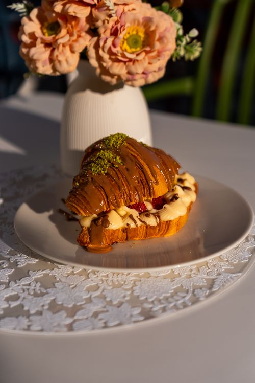 Free Delicious chocolate-drizzled croissant with cream, garnished with strawberries and pistachios, beside a floral display. Stock Photo