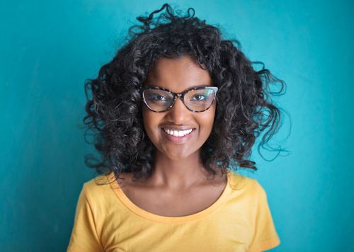 Free Young happy black lady with curly hair in eyeglasses and casual outfit toothy smiling while standing against blue background and looking at camera Stock Photo