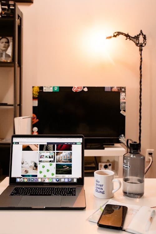 Desktop computer and open netbook with images on screen near mug of coffee with bottle of water and diary with smartphone on table behind glowing lamp in apartment