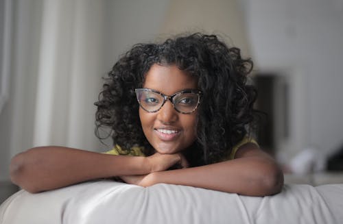 Cheerful young African American female with curly hair and eyeglasses lying on comfortable sofa with hands under chin and smiling while looking at camera