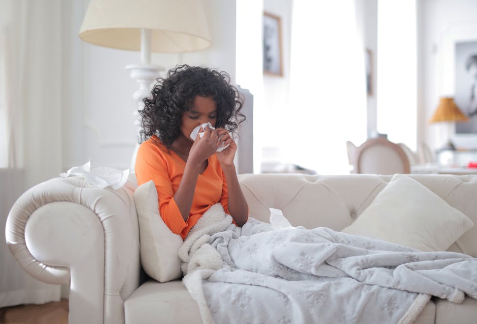 Side view of sick African American female blowing nose while sitting on sofa under blanket in cozy modern home in daylight