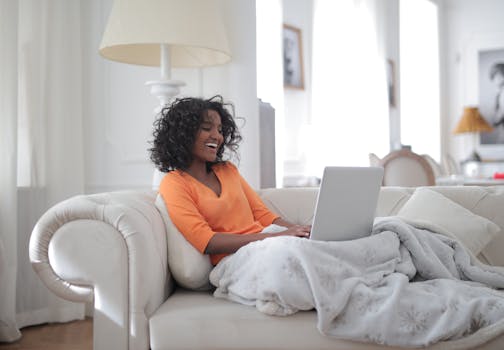 A cheerful woman uses her laptop, relaxing on a cozy couch at home.