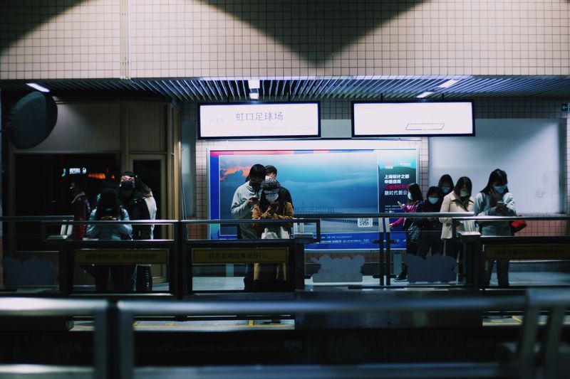 Passengers stand in line at a dimly lit subway station, focused on their devices.