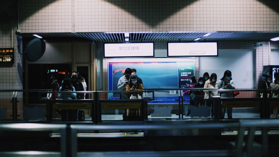 Passengers stand in line at a dimly lit subway station, focused on their devices.