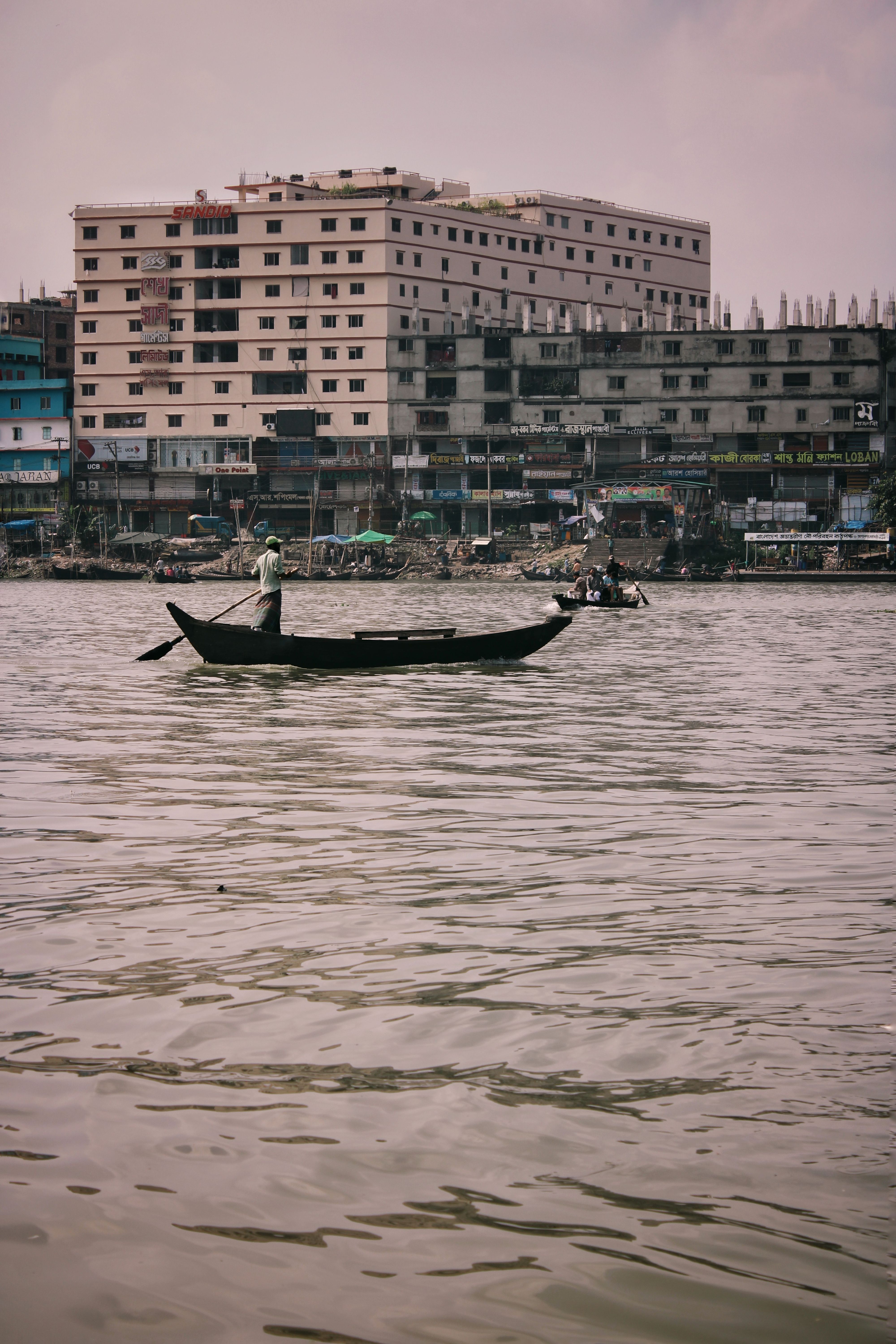 A traditional wooden boat with a rower on a river against the backdrop of Dhaka's urban waterfront.