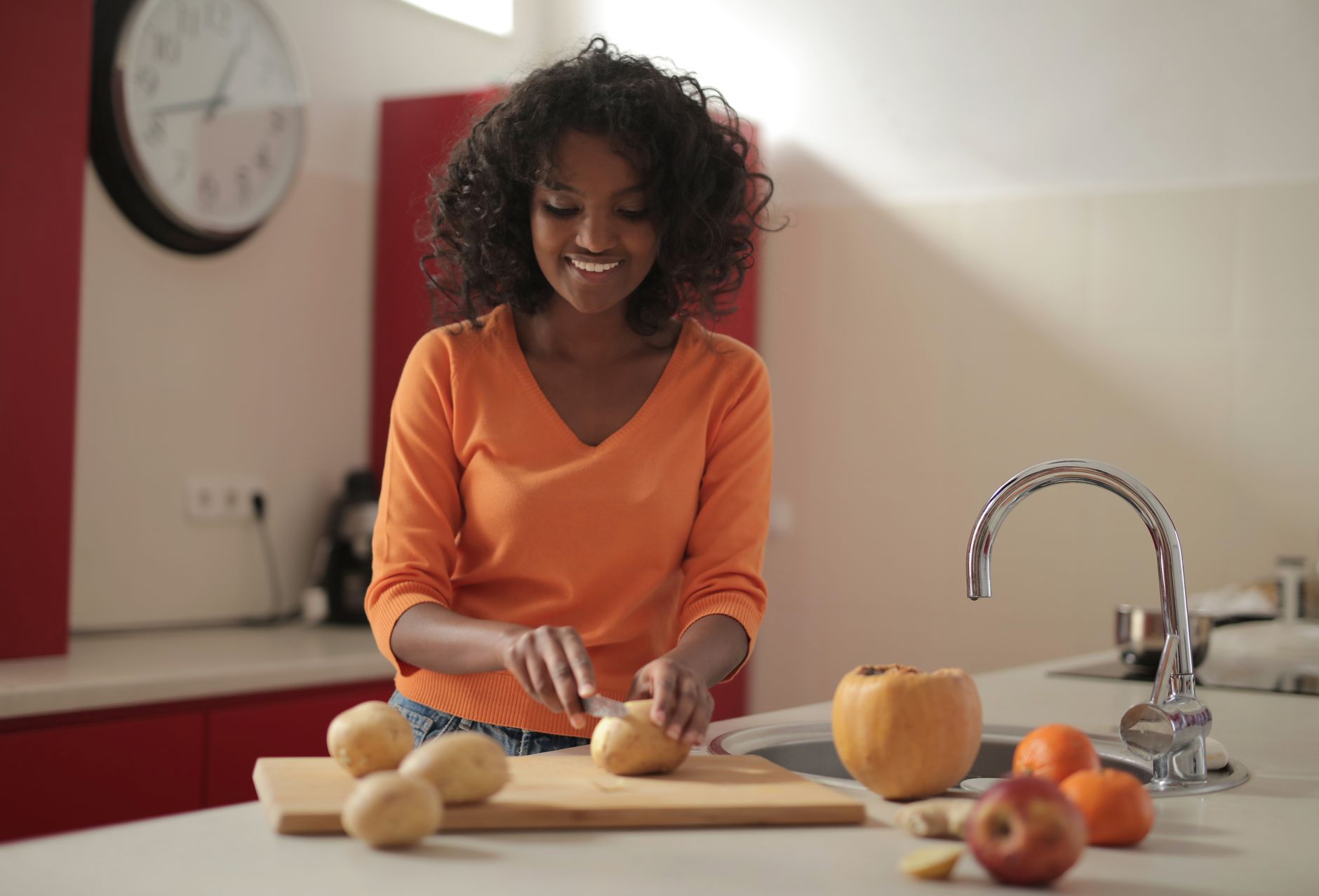 Happy African American woman in casual wear smiling while cutting potatoes on board and fruits placed aside on countertop in cozy kitchen in daylight