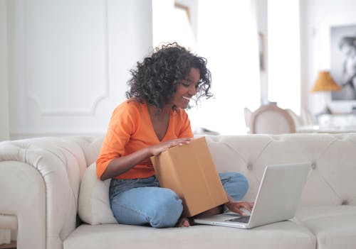 Free Side view of smiling African American lady in casual clothes holding box while sitting on sofa and browsing laptop Stock Photo