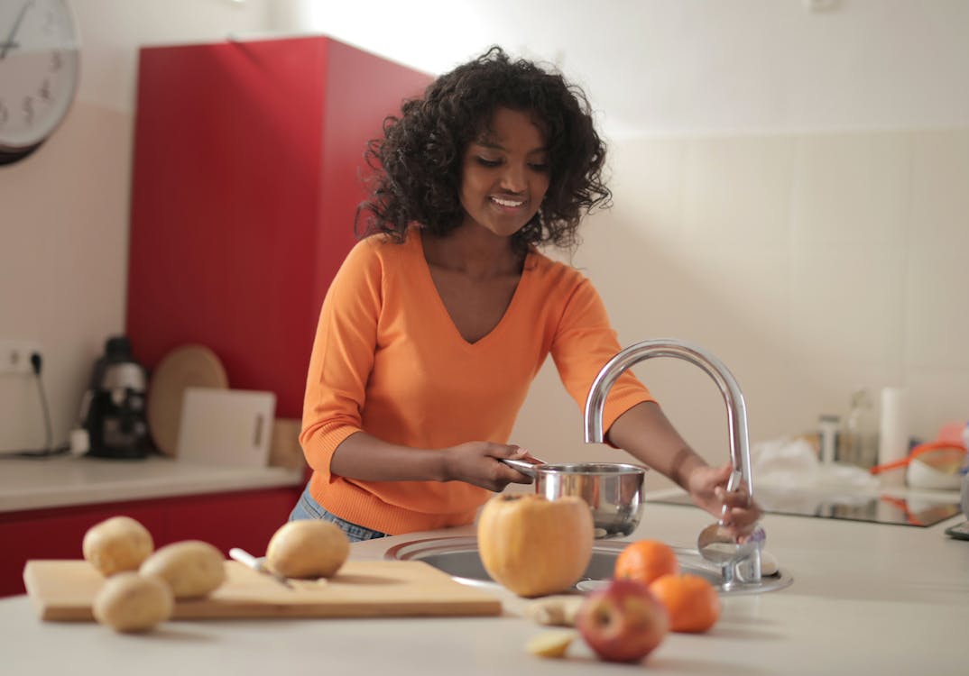 Free Smiling young ethnic lady in colorful casual clothes standing near sink and pouring water in pot while cooking food in spacious modern kitchen at home Stock Photo