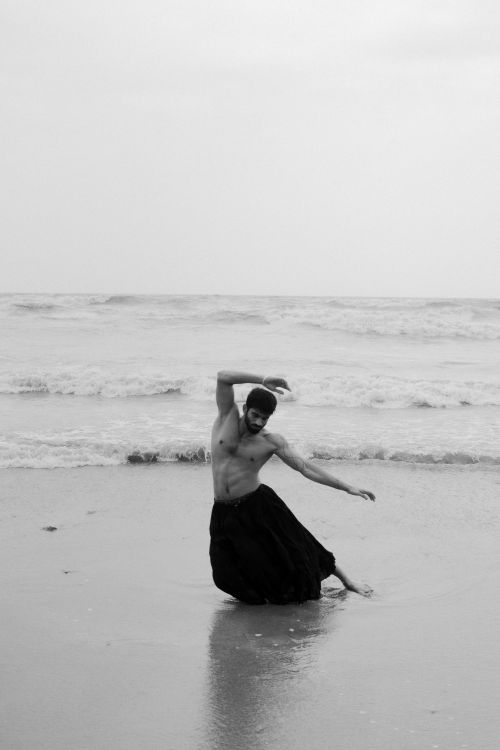 Free A shirtless dancer gracefully performs on a serene beach under an overcast sky. Stock Photo