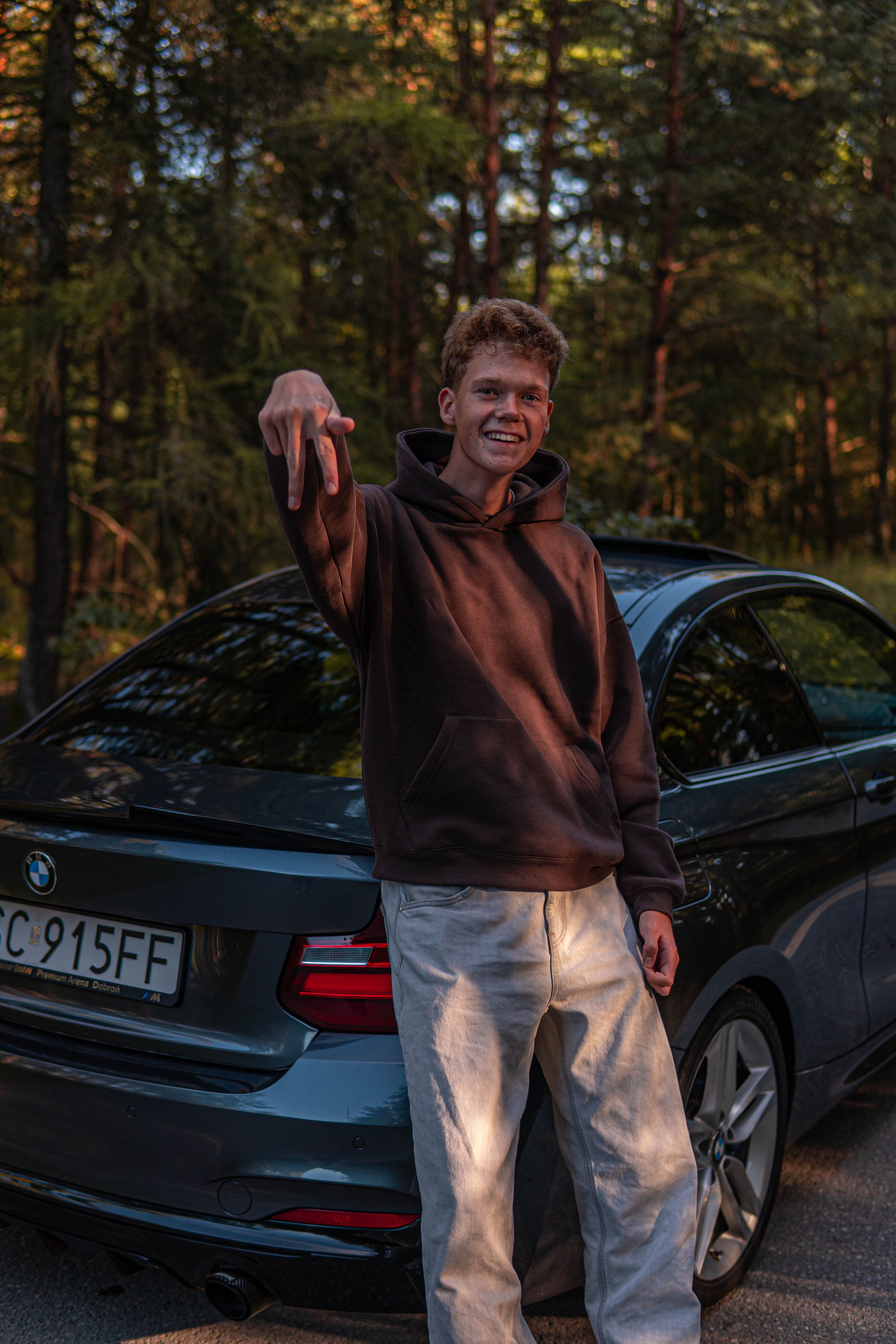 Smiling young man in hoodie stands by a black BMW in a scenic forest setting during the day.