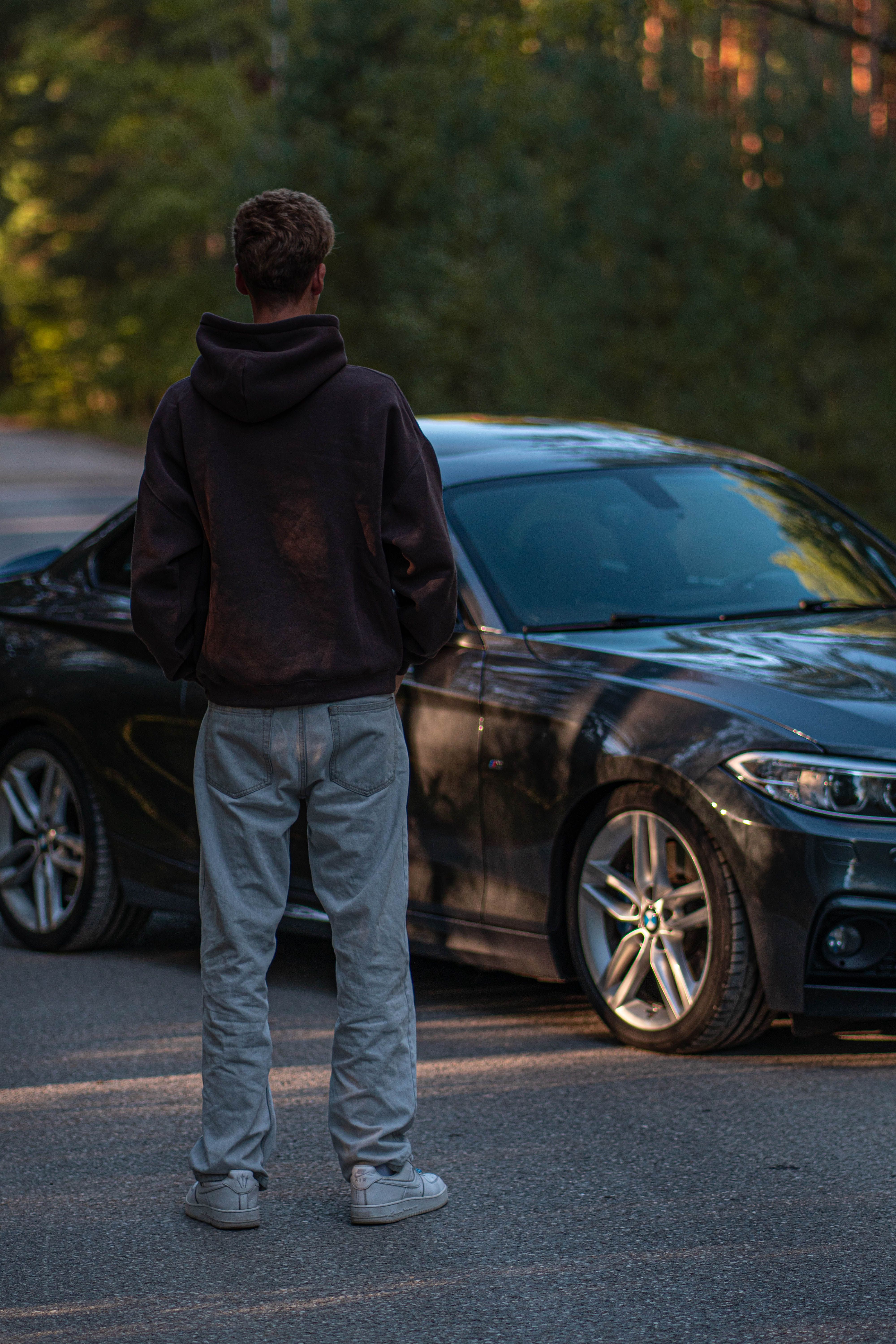 A young man in casual attire stands next to a sleek luxury car parked on an outdoor road.