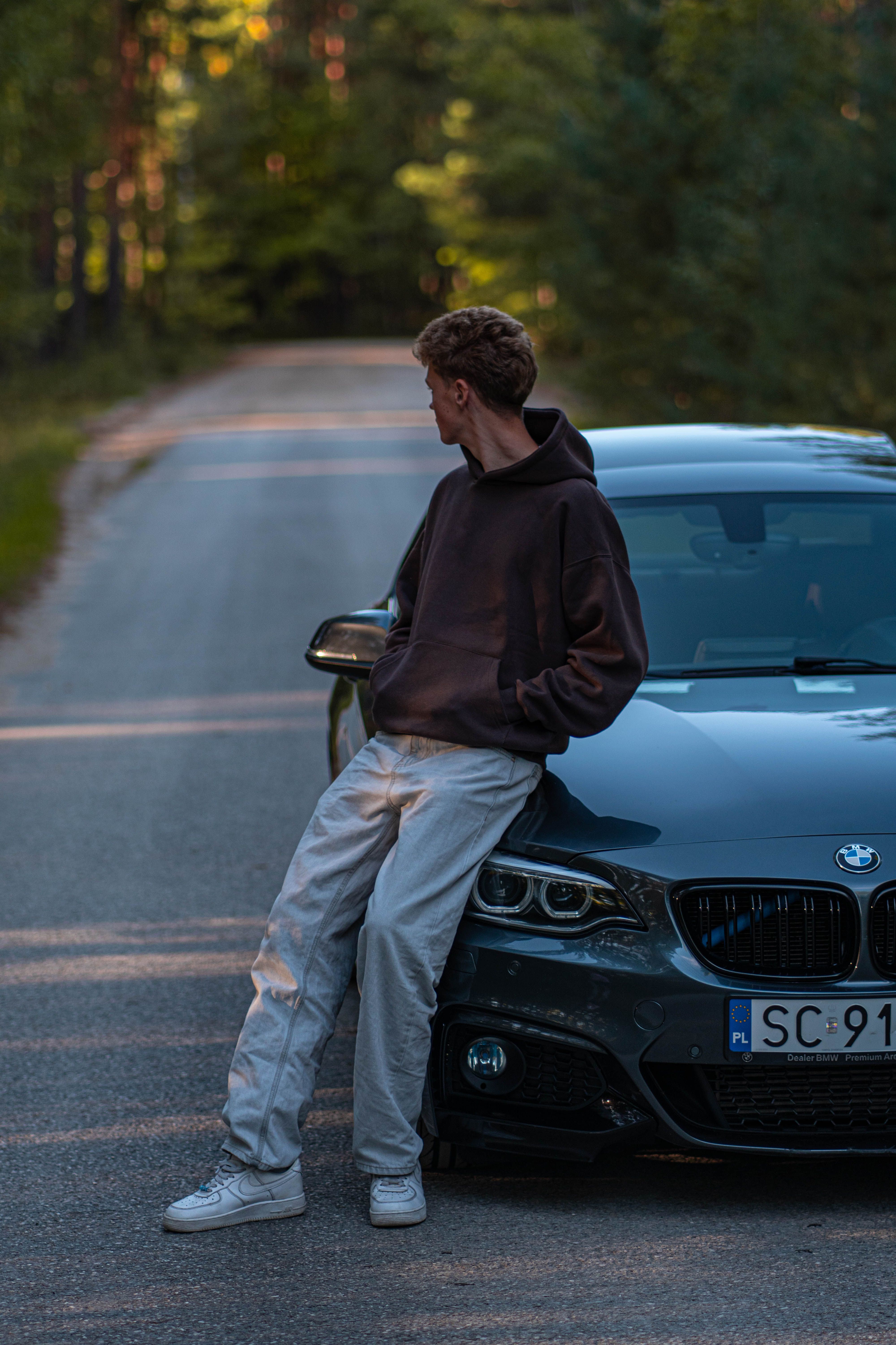 A young man leans on a BMW car parked on a forest road, showcasing a serene outdoor scene.