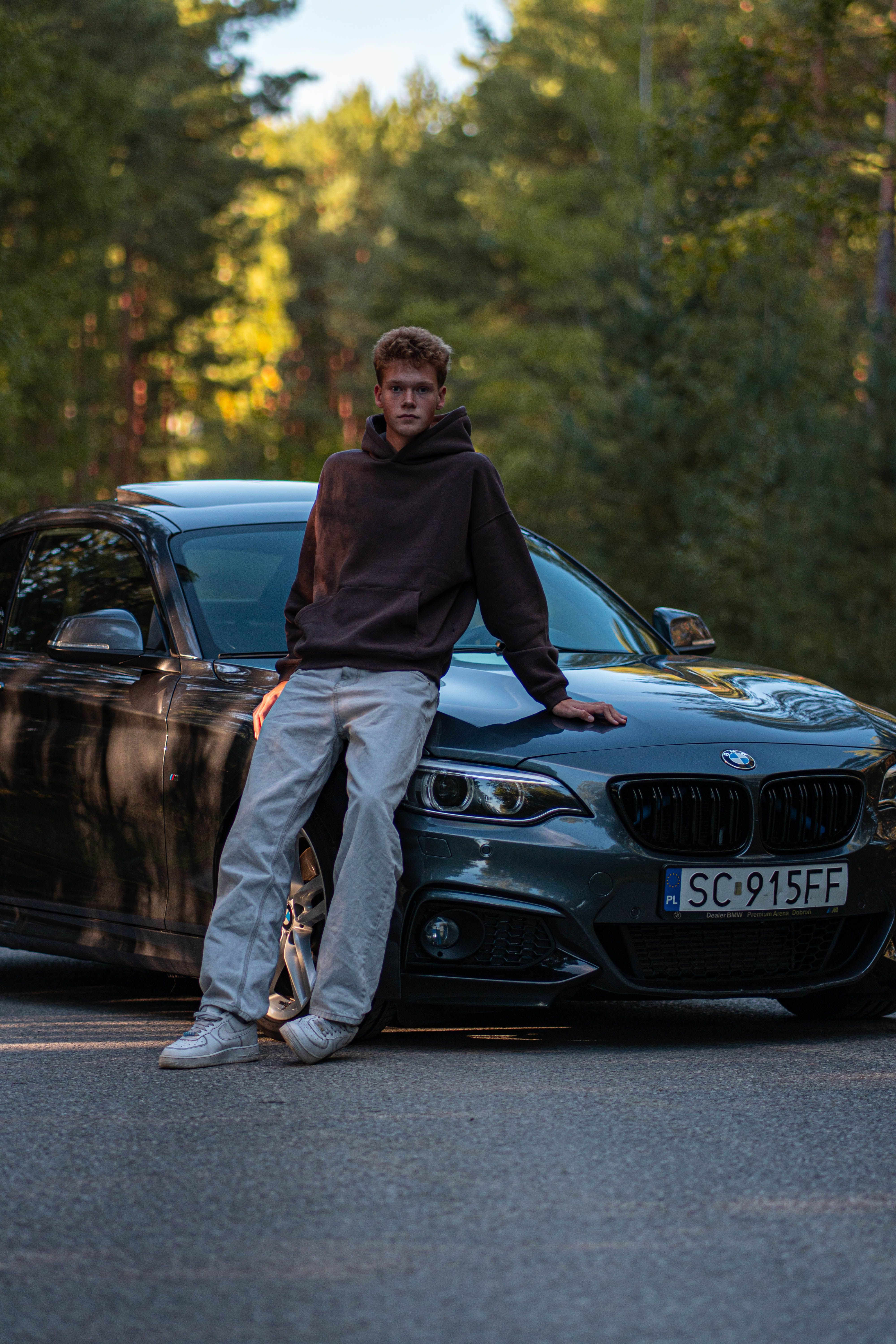 A young man leans on a BMW car surrounded by trees in a serene outdoor setting.