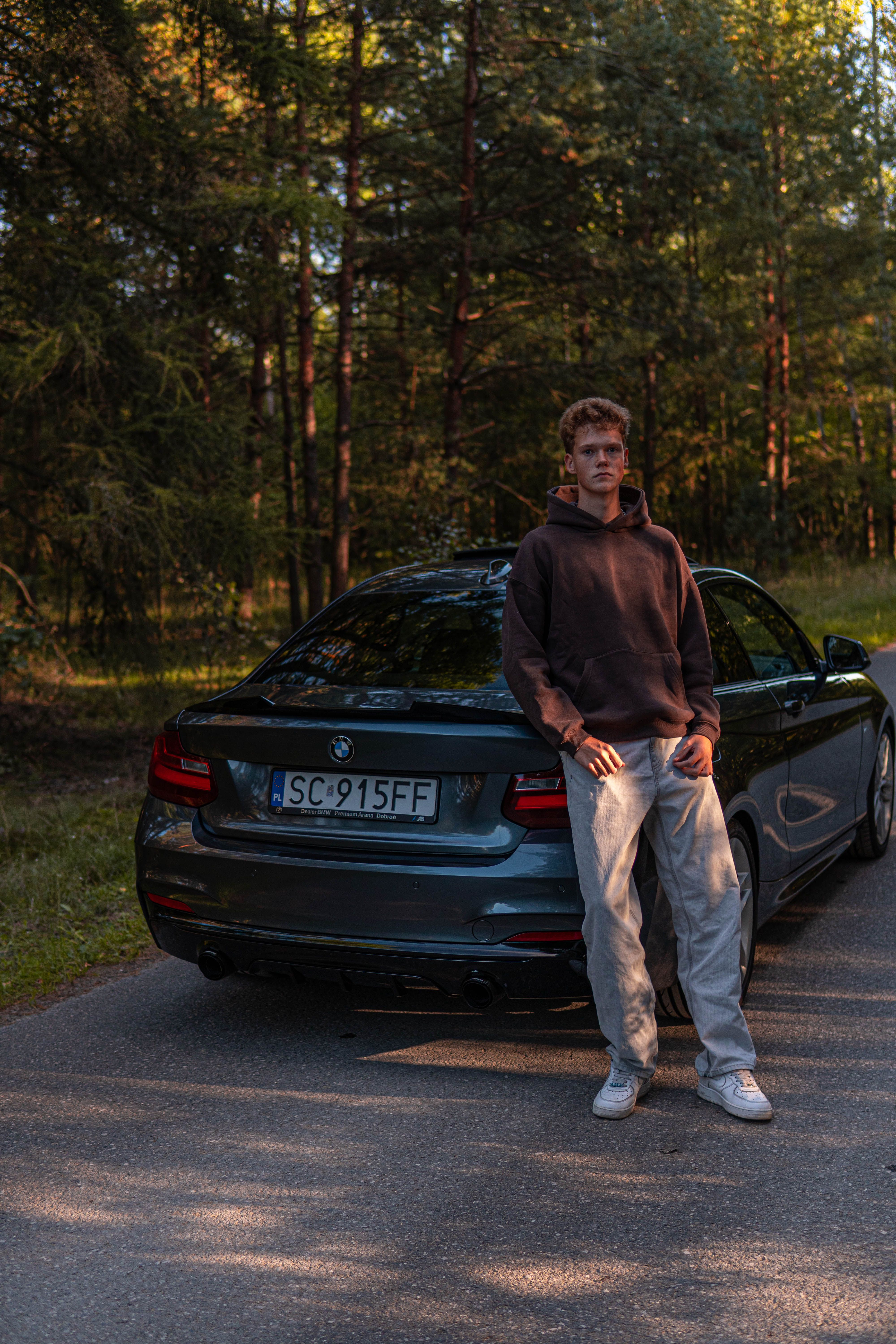 Casual portrait of a young man standing next to a BMW on a scenic forest road at sunset.