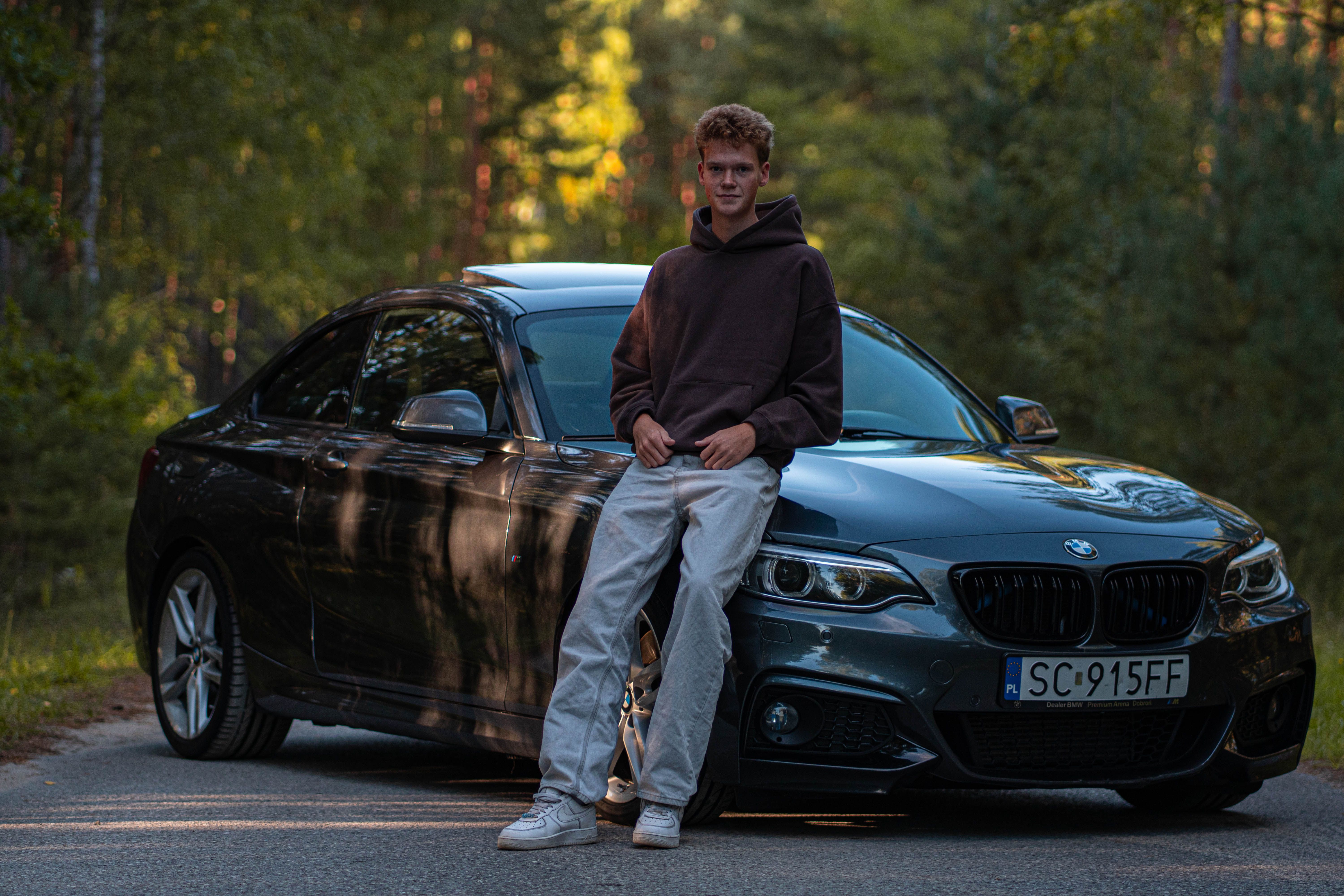 A young man leans on his BMW car in a serene forest environment, evoking a sense of adventure.