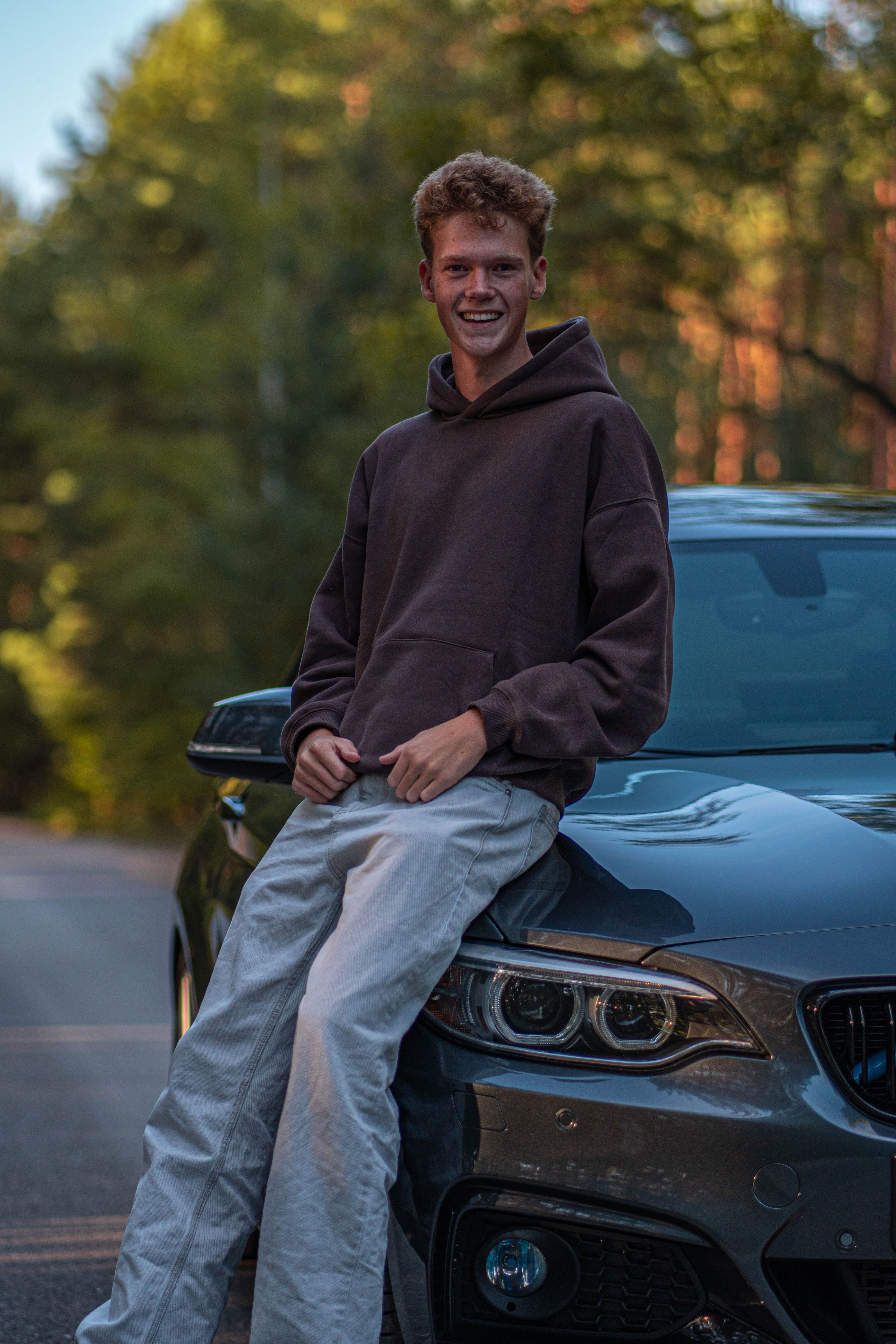 Smiling young man leaning on a car in a forest setting, enjoying a casual moment.