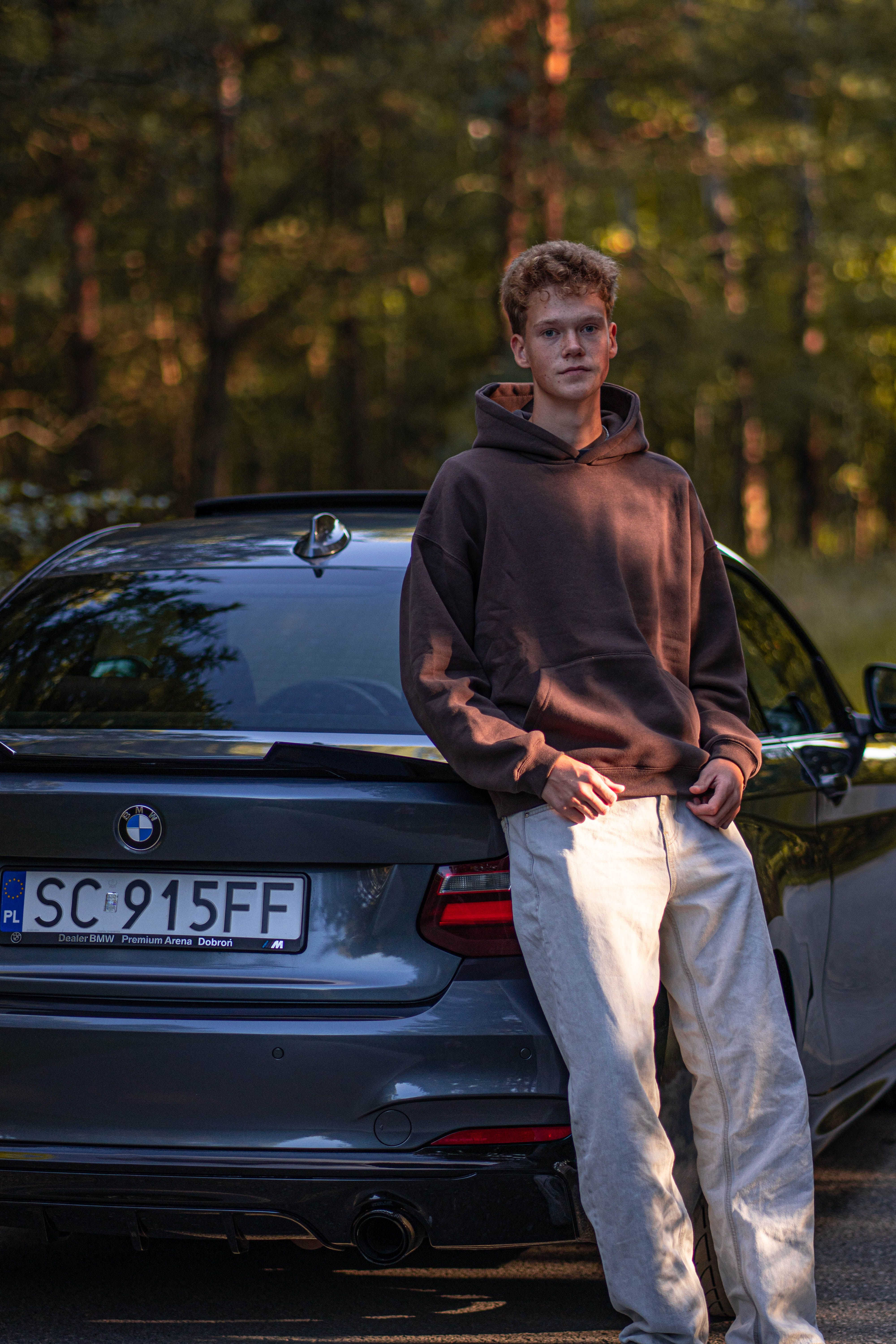A young man leans against a black sporty car amidst a serene forest ambiance.