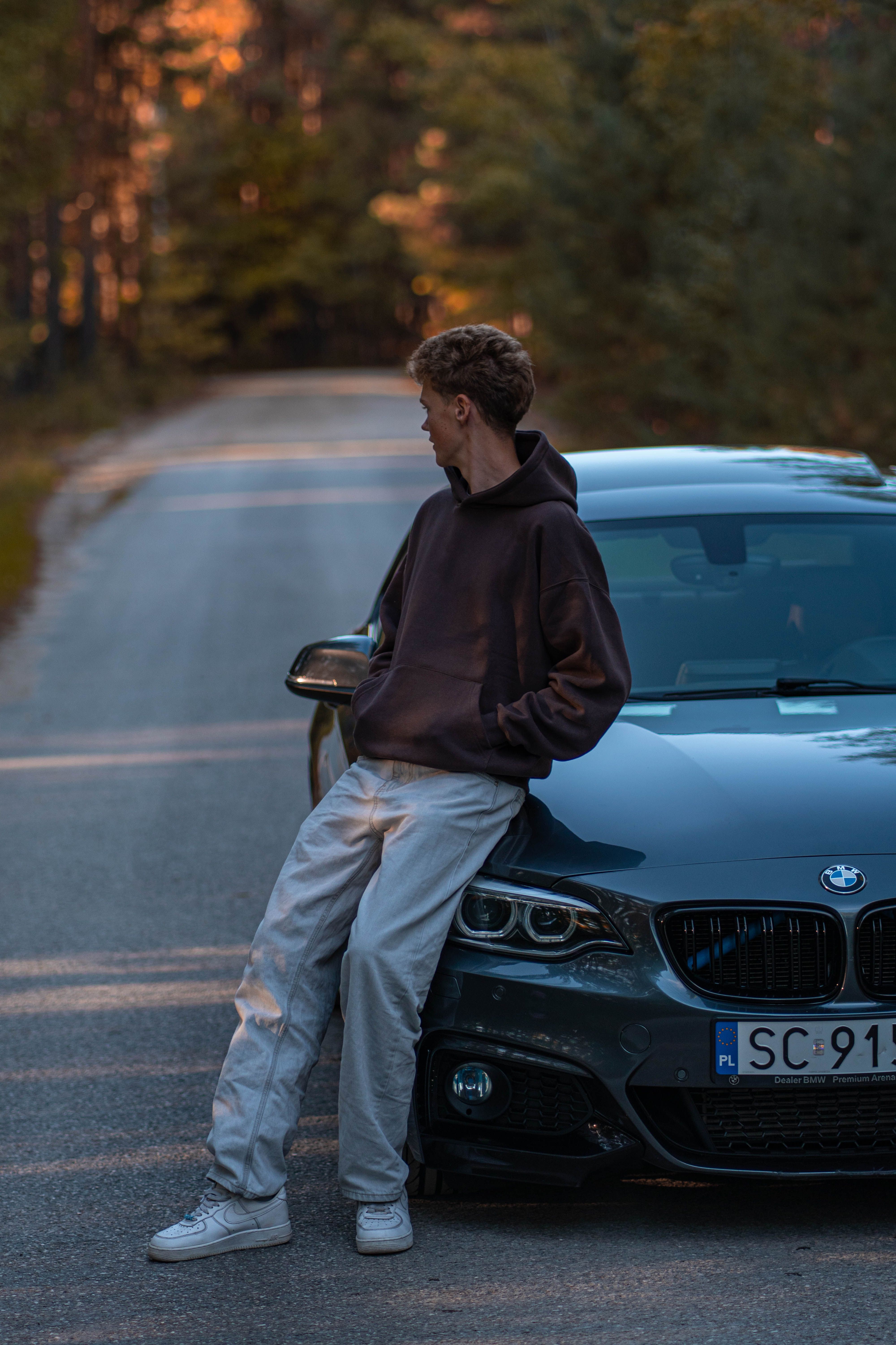 Young adult casually leaning on a BMW M car parked on a forest road during golden hour.