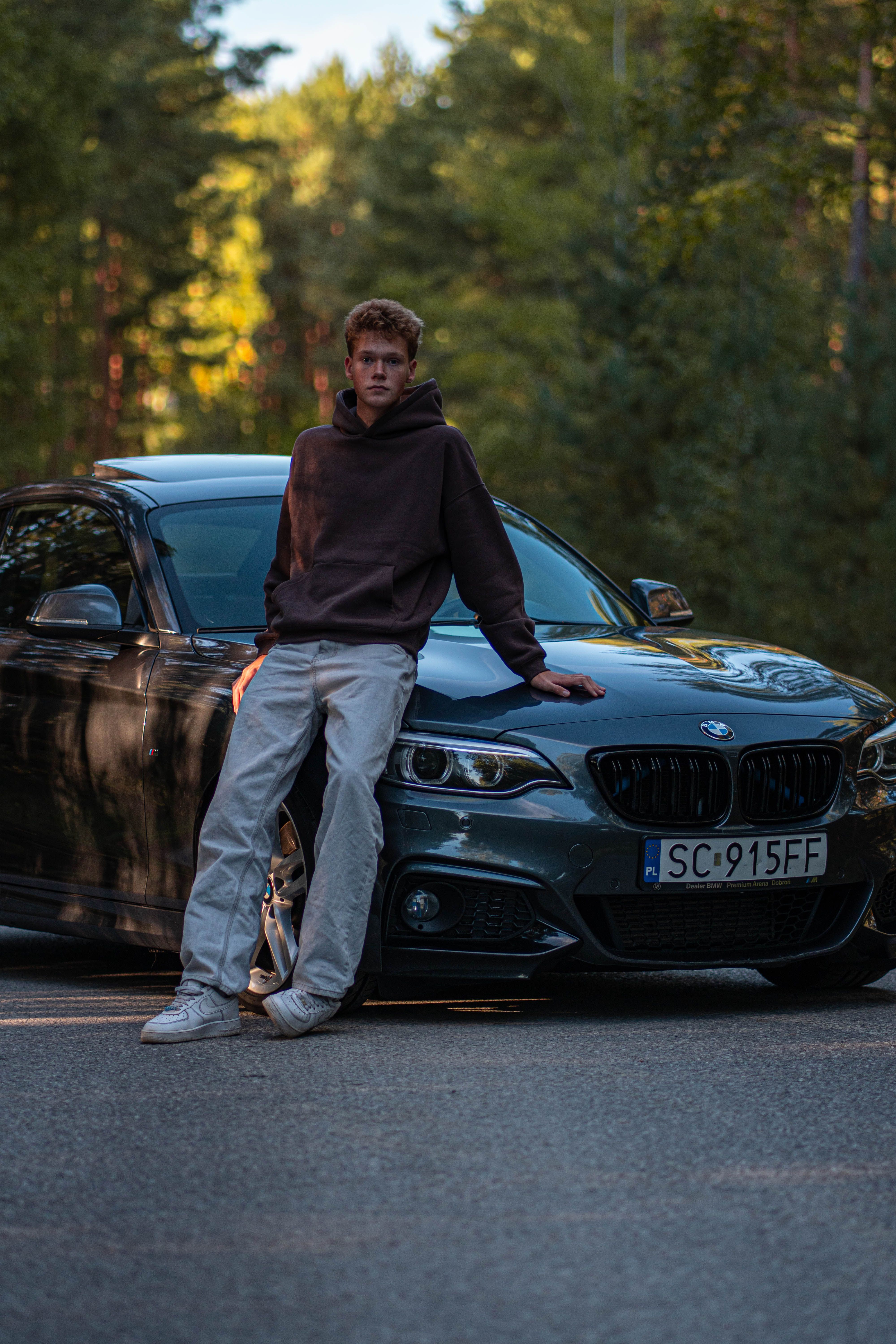 Teen with a hoodie leaning on a BMW on a sunlit road through a dense forest.