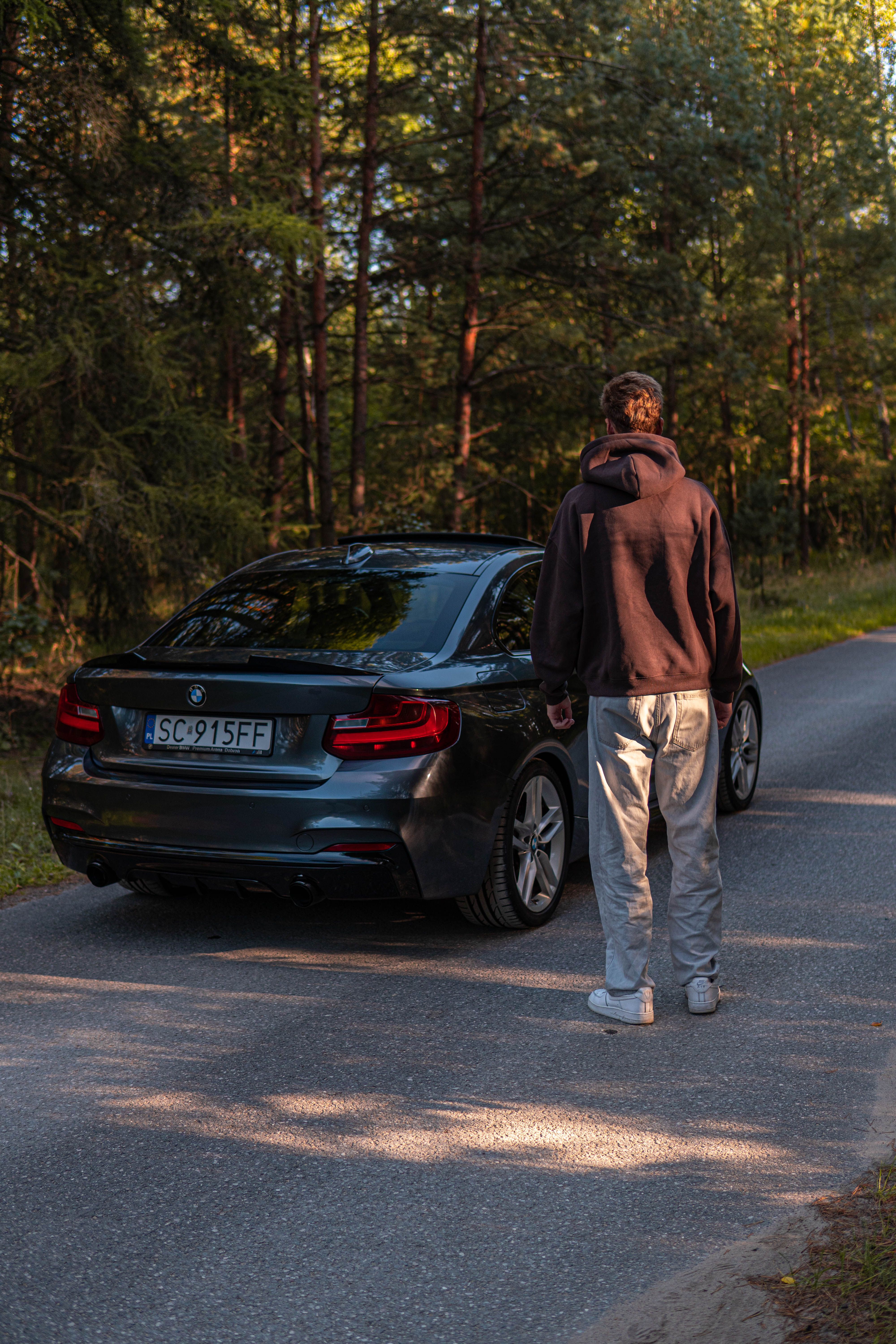 A person stands beside a parked BMW on a quiet forest road in daylight.