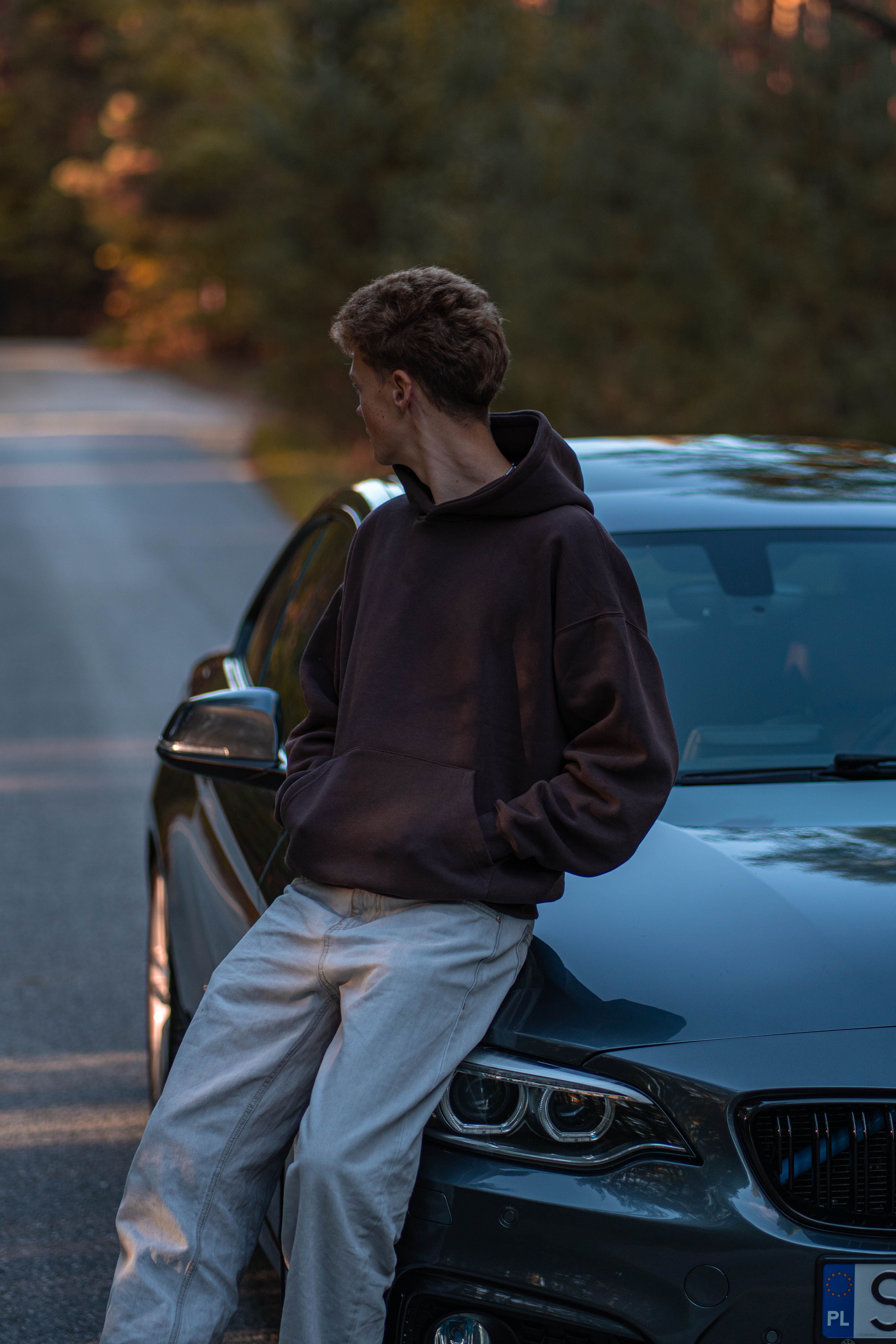 A young man in casual attire leans on a sleek sports car parked on a serene forest road.