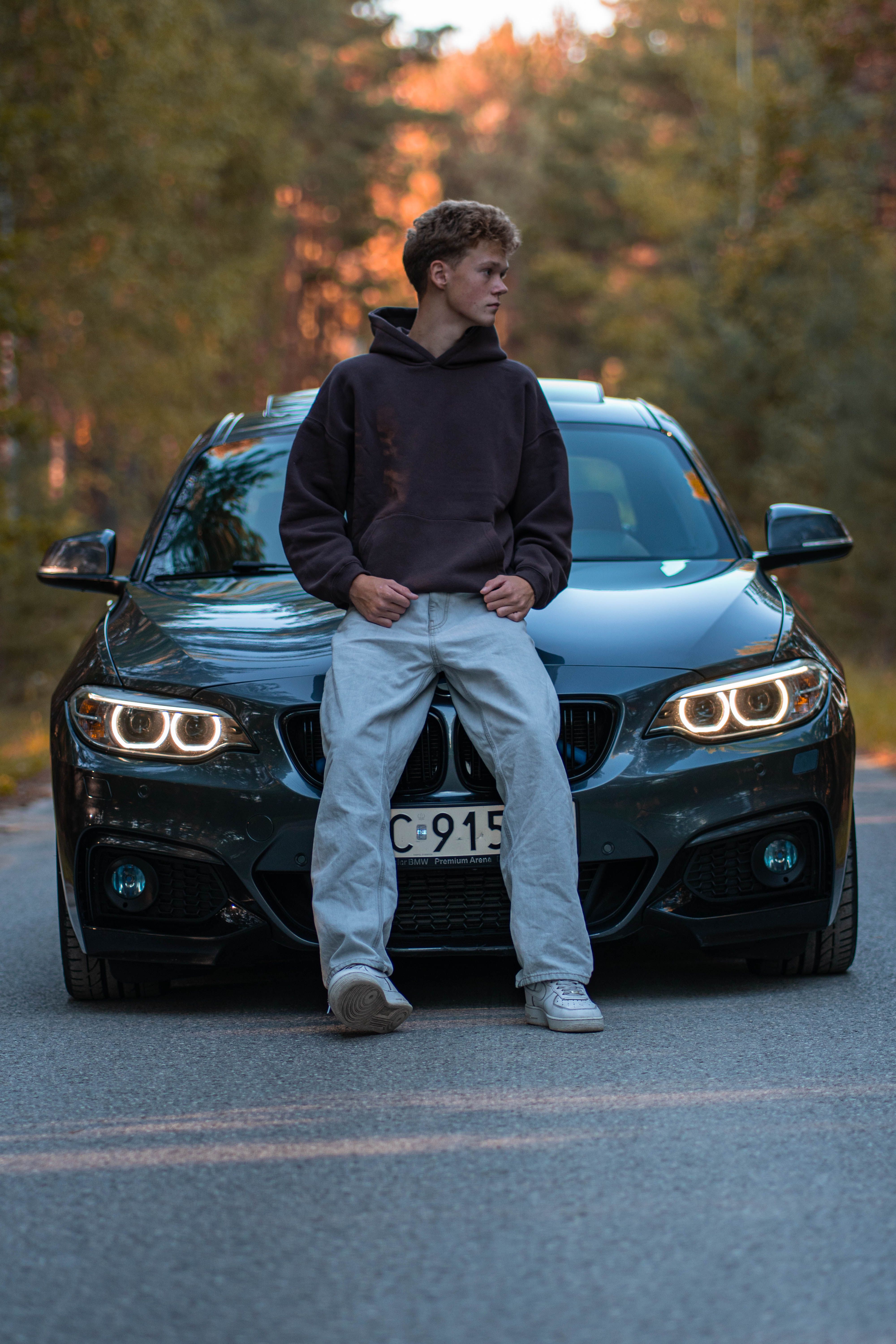 A young man confidently leans on a sleek car on a scenic road.