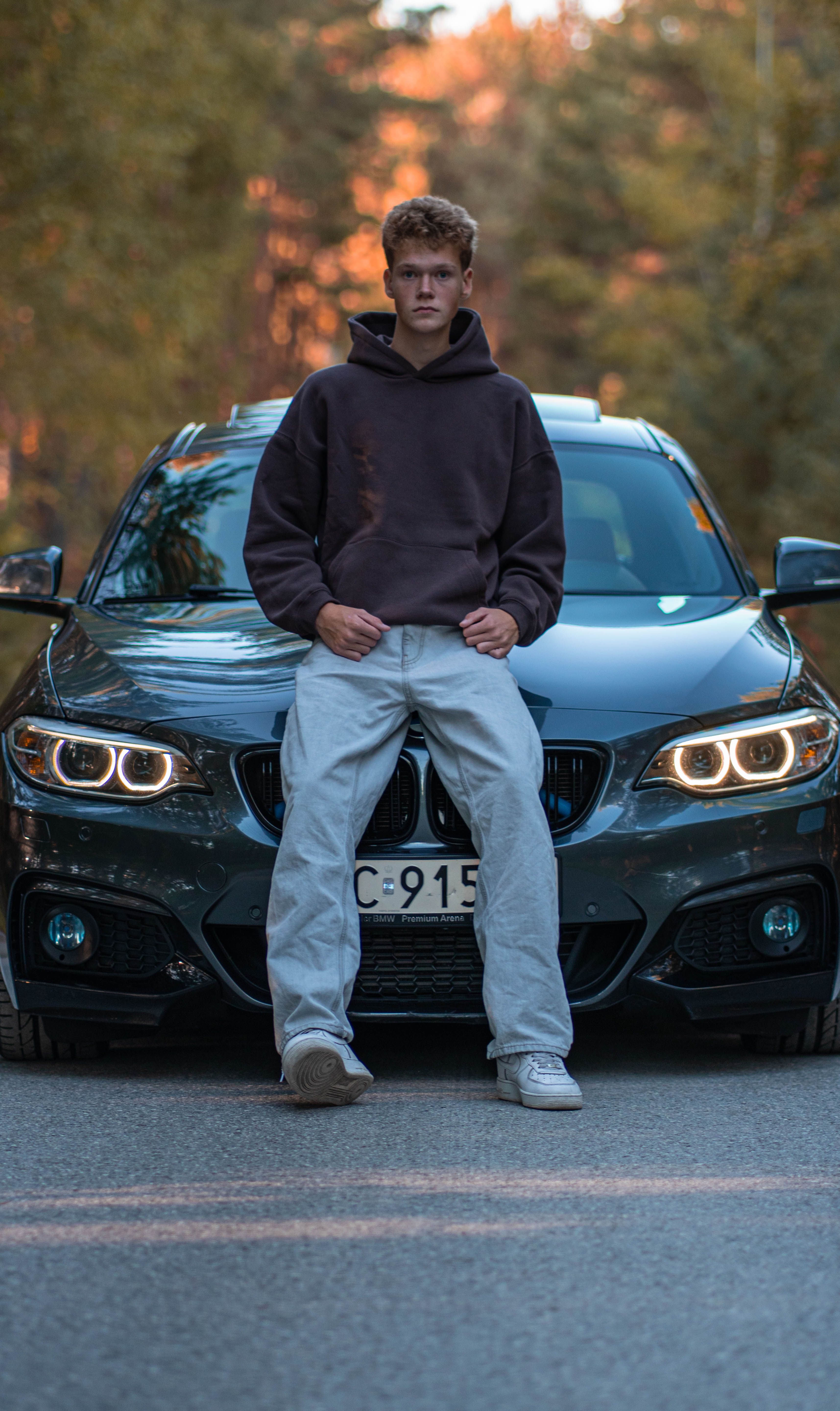 A young man leans against a sleek BMW in a serene forest setting, showcasing style and luxury.