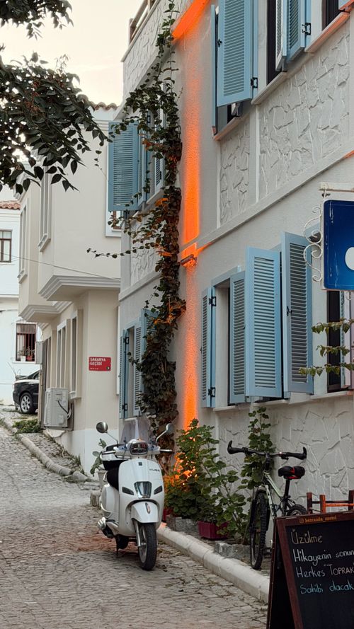 Free A quaint alley in Bozcaada, Türkiye, featuring a Vespa scooter, bicycle, and charming architectural elements with blue shutters. Stock Photo