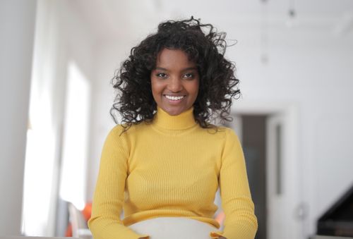 Free Cheerful young ethnic lady in casual yellow sweater sitting on comfortable seat in light living room and smiling while looking at camera Stock Photo