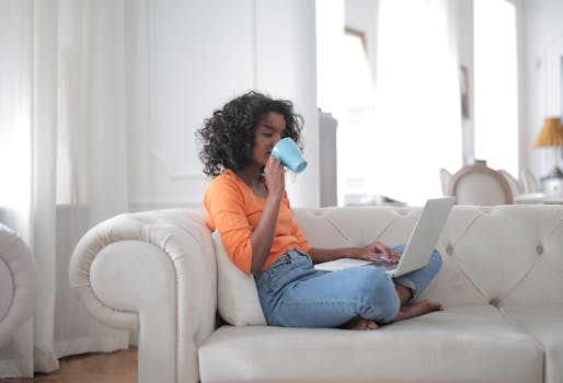 Side view of positive young ethnic lady in casual sweater and jeans drinking coffee while working on laptop on comfortable sofa in light living room at home