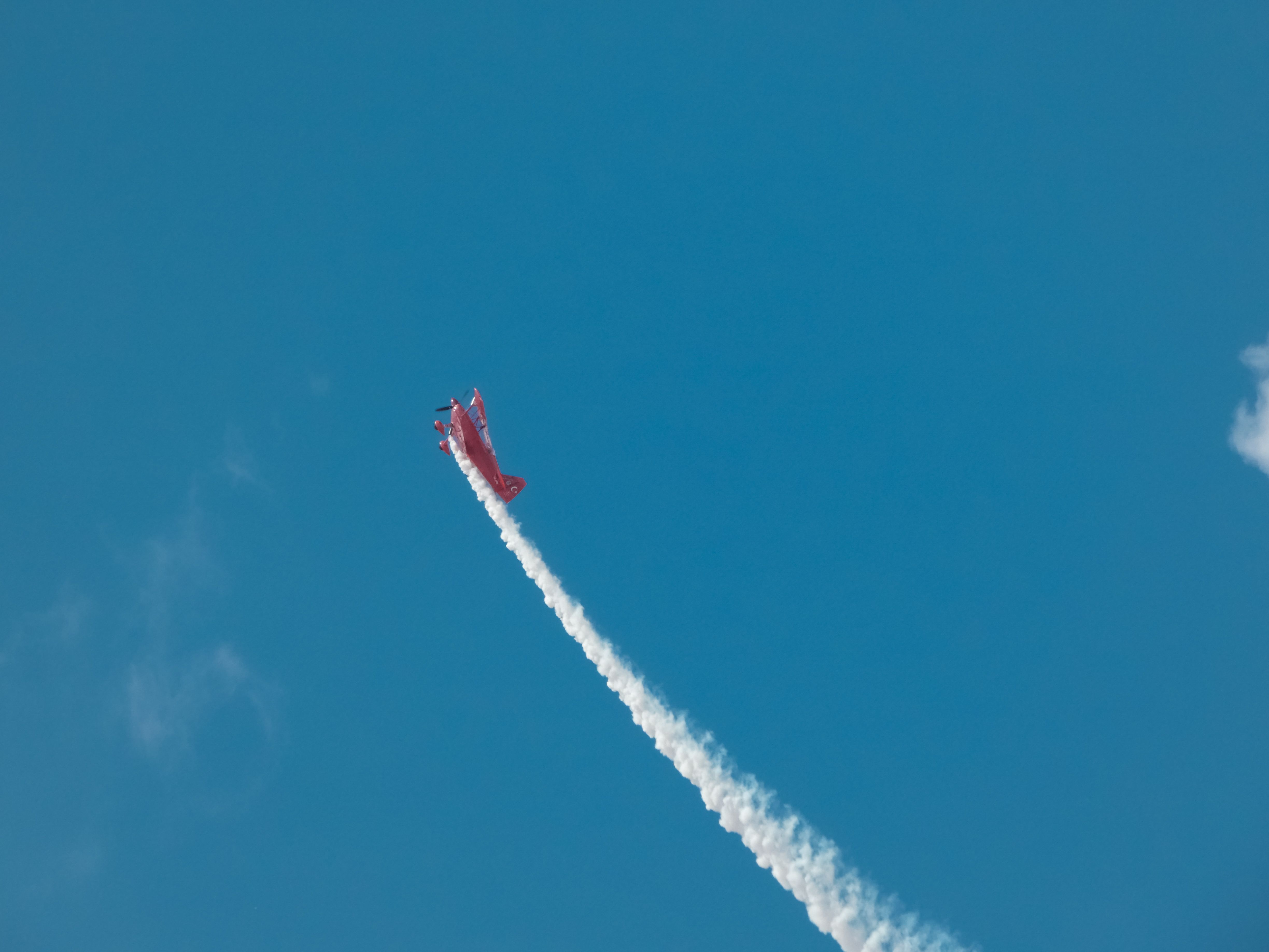 A red aerobatic plane soaring in a clear blue sky, trailing white smoke during an air show.