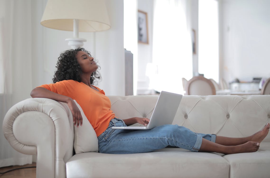 Woman in Orange Top and Blue Denim Jeans Sitting on White Sofa Using Macbook
