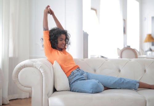 Free Woman in Orange Top and Blue Denim Jeans Sitting on White Couch Stock Photo