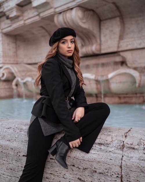 Woman in Black Coat and Black Pants Sitting on Concrete Near Water Fountain