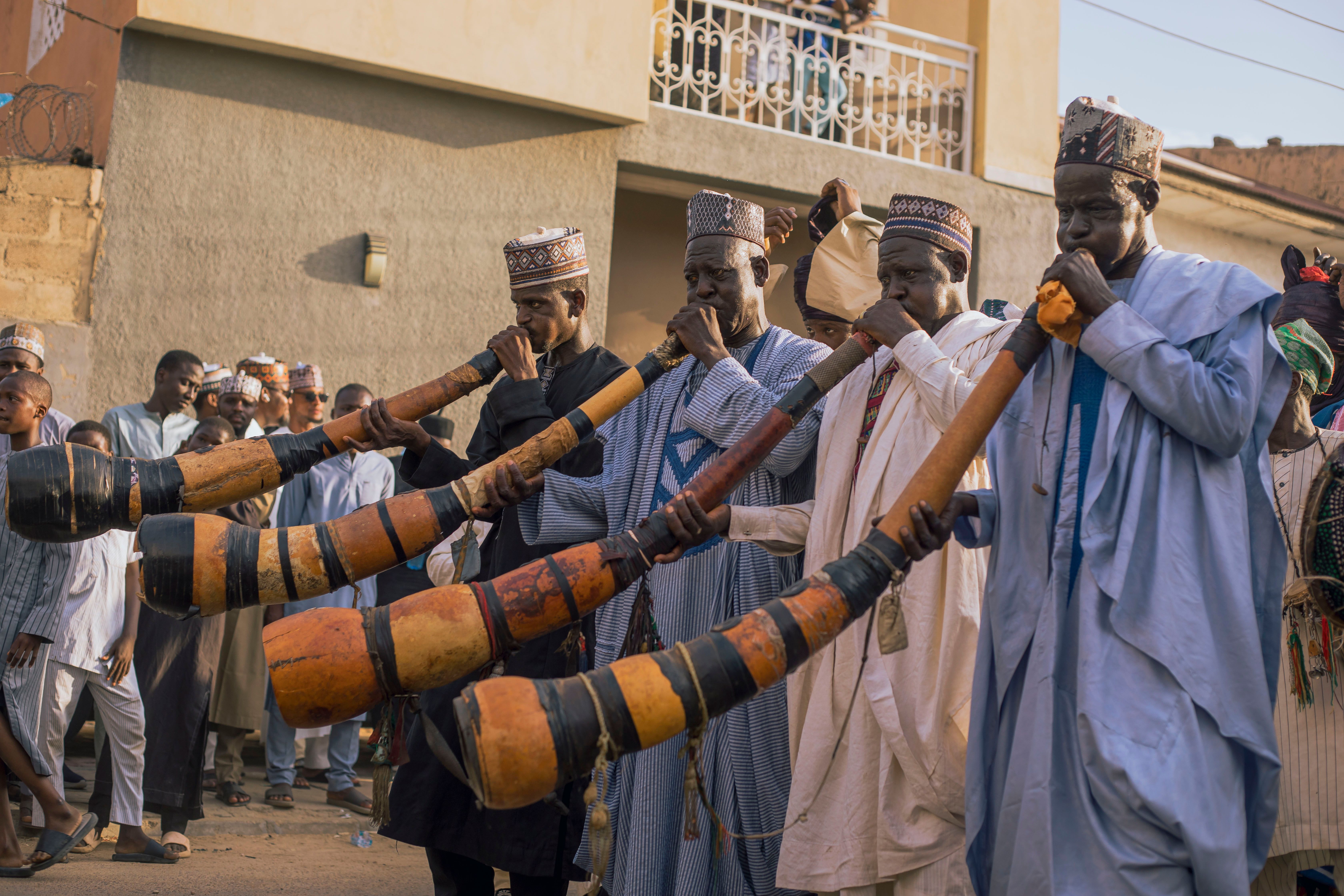 A group of Nigerian men in traditional attire playing long wooden horns during a cultural event outdoors.