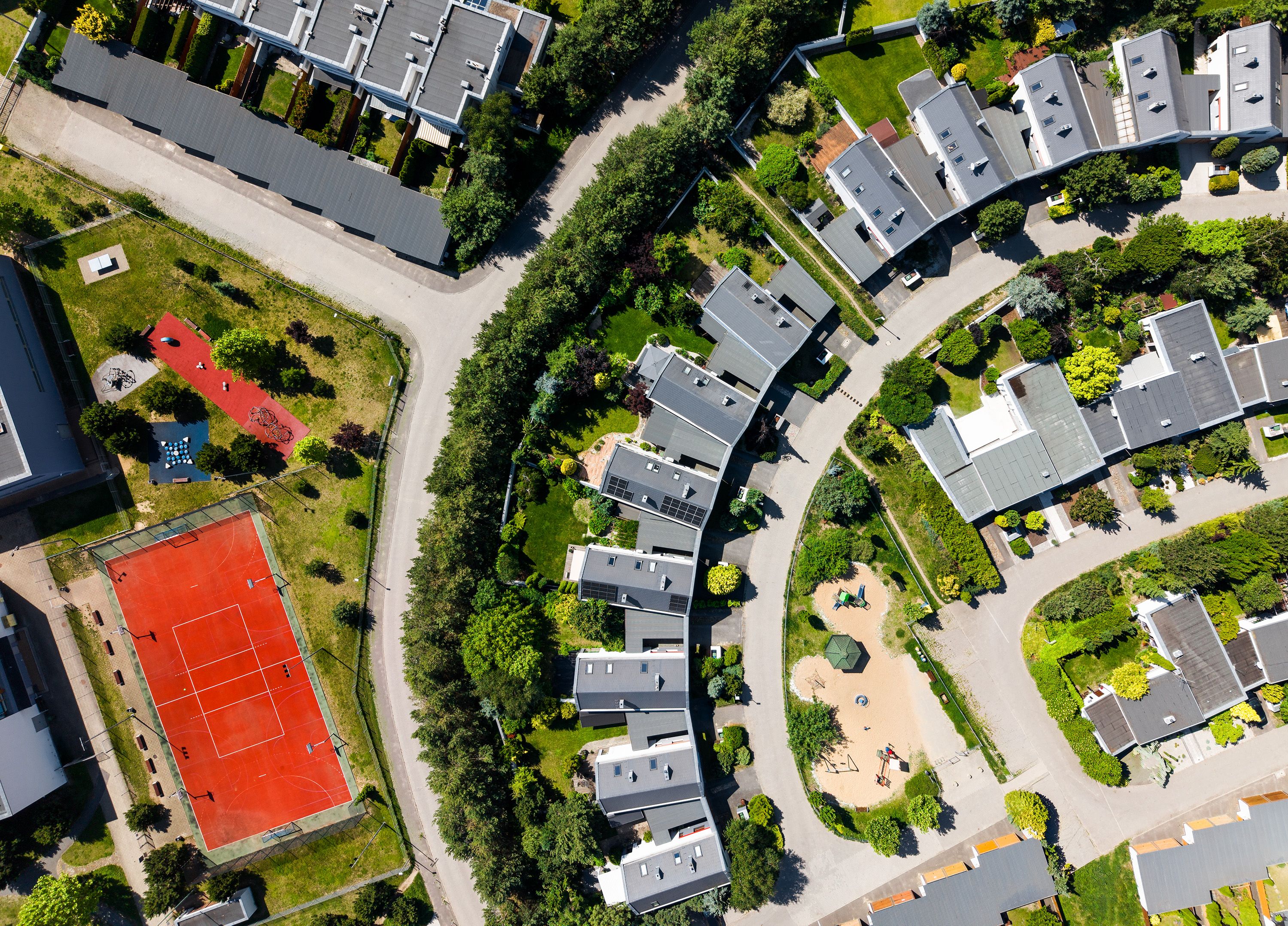 Drone shot of a modern residential area with houses, greenery, and a red tennis court on a sunny day.
