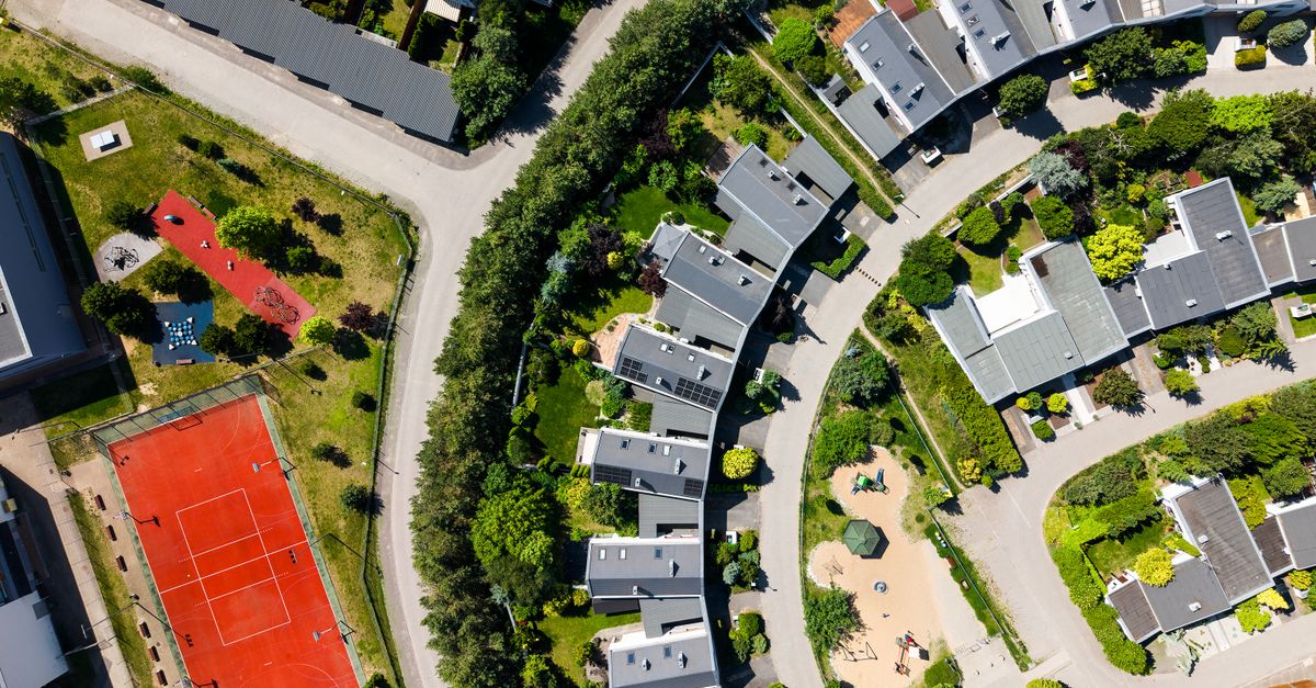 Drone shot of a modern residential area with houses, greenery, and a red tennis court on a sunny day.
