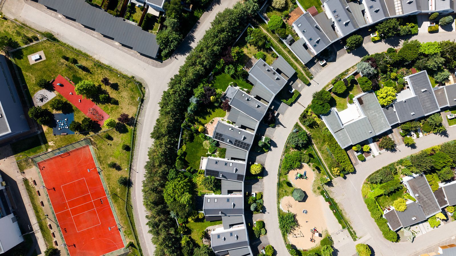 Drone shot of a modern residential area with houses, greenery, and a red tennis court on a sunny day.