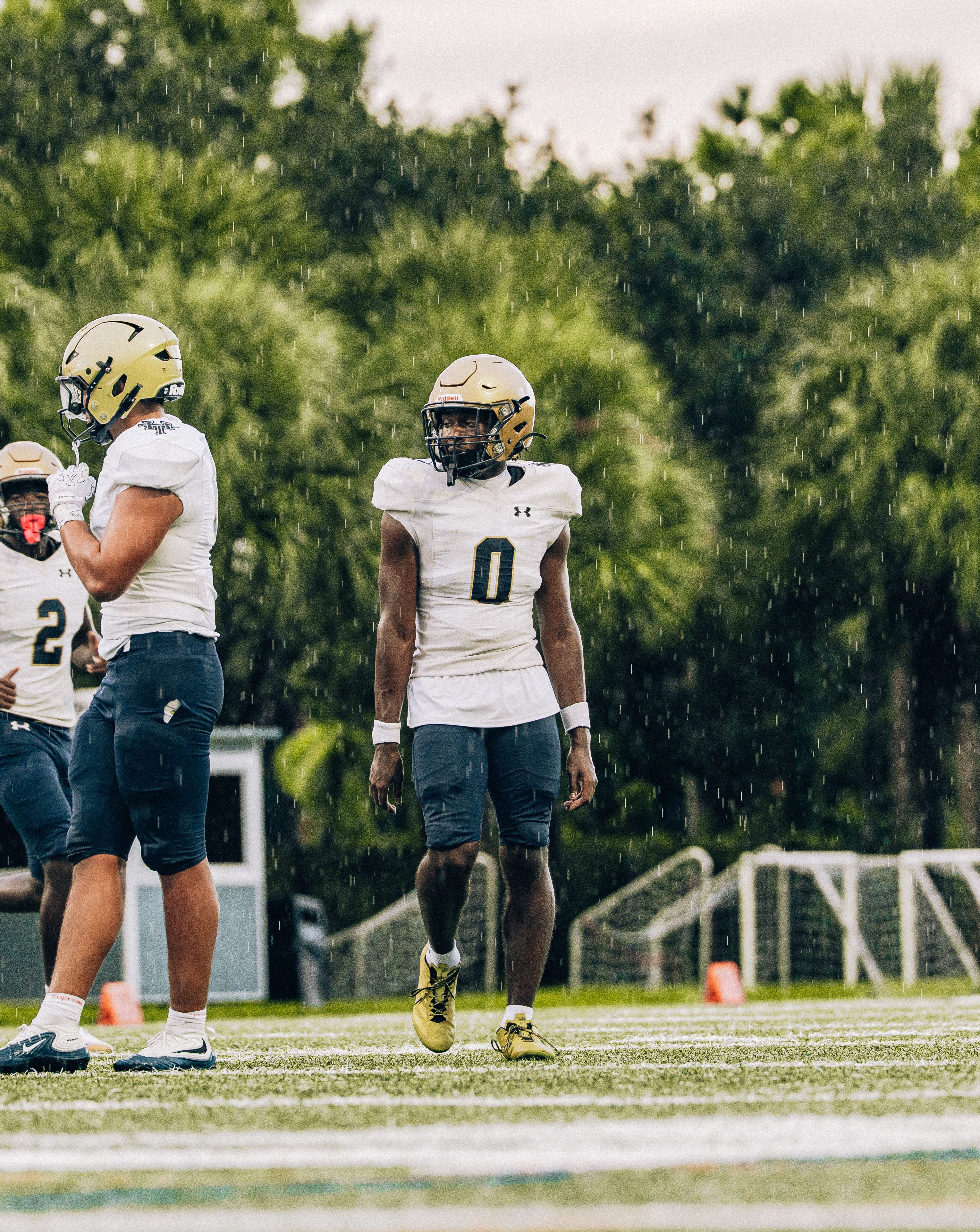 Football players on field during a rainy day, preparing for a game under lush greenery.