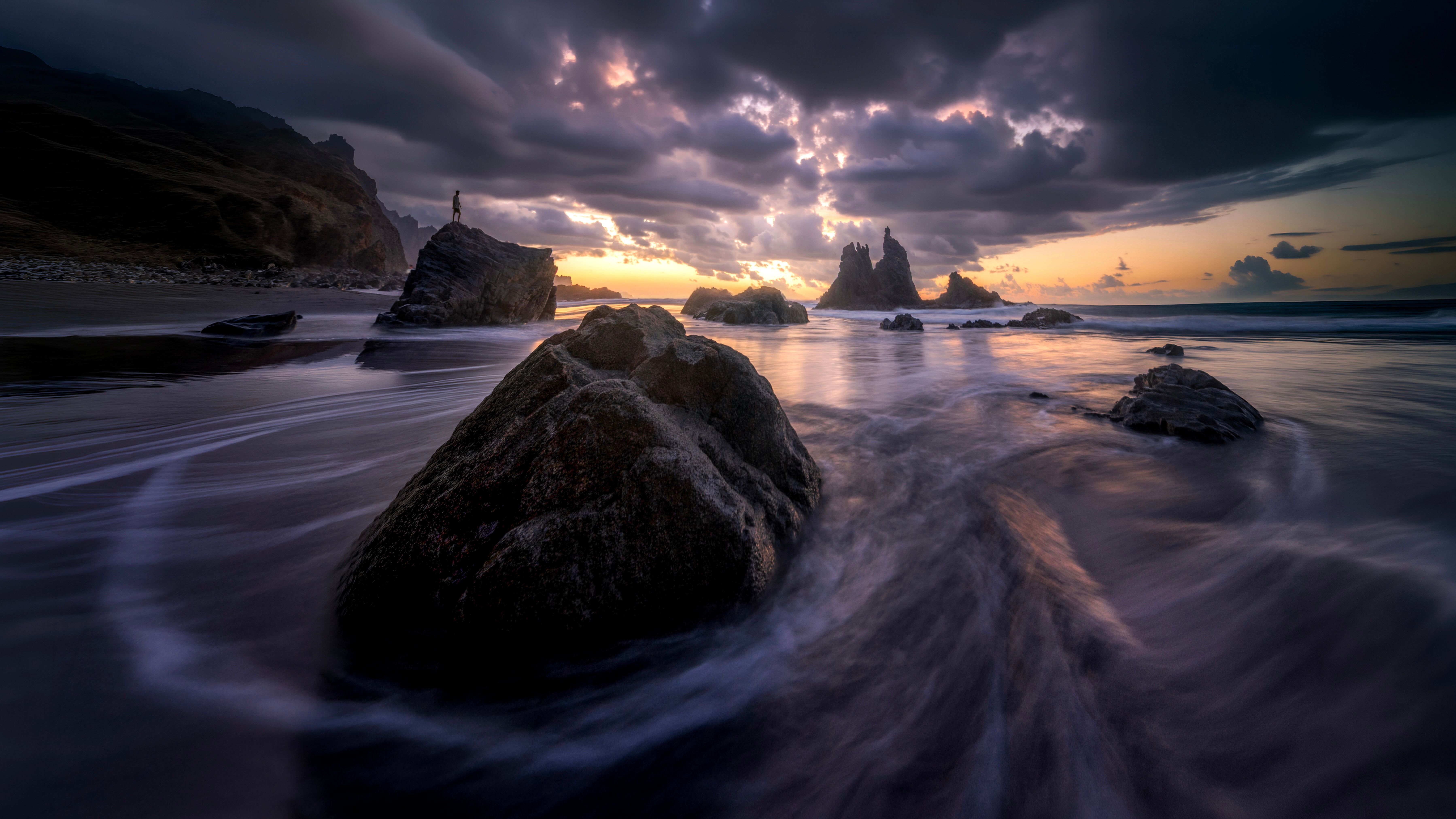 Dynamic seascape at sunset featuring rocks and a silhouetted figure by the water's edge.