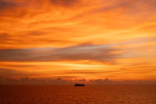 Free A cargo ship silhouetted against a fiery orange sunset over a calm sea. Stock Photo
