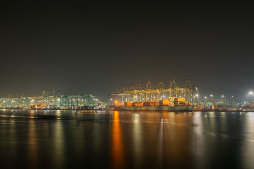 Free A bustling container port at night, featuring illuminated cranes and cargo ships reflecting on calm waters. Stock Photo